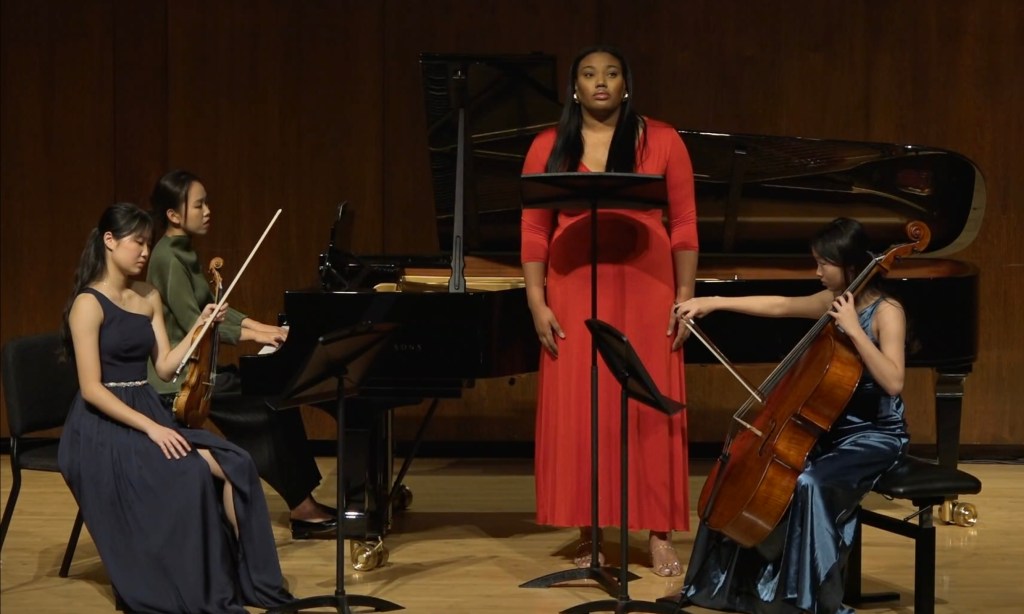A performance scene featuring four musicians: a singer in a red dress stands in front of a music stand, while three instrumentalists, including a pianist at a grand piano, a violinist, and a cellist, are seated, contributing to the musical ensemble.